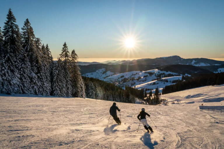 Zwei Personen fahren bei Sonnenuntergang auf einer verschneiten Piste bergab, umgeben von schneebedeckten Bäumen und fernen Bergen unter einem klaren Himmel in der Nähe des Hotels Hochfirst.