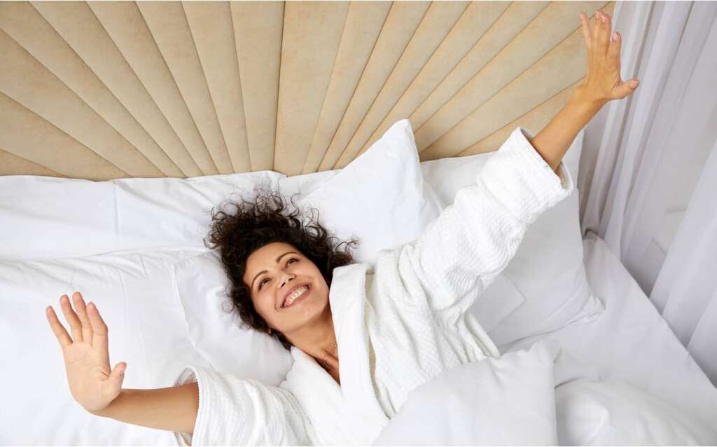 Lächelnde Frau wacht im Bett auf und streckt die Arme nach oben