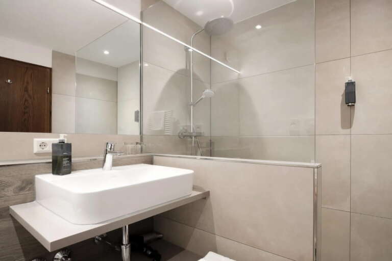 Modernes Badezimmer im Hotel Hochfirst mit weißem, rechteckigem Waschbecken, Wandarmatur, großem Spiegel, beige gefliesten Wänden und einer begehbaren Dusche mit Glaswand.