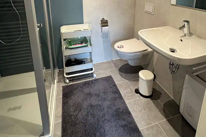Modernes Badezimmer mit Dusche, wandhängendem WC, Waschbecken, Rollwagen mit Toilettenartikeln, kleinem Mülleimer und einer dunkelgrauen Fußmatte.