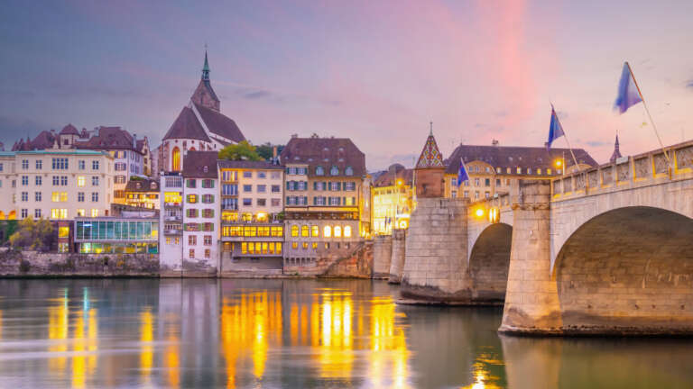 Basel am Flussufer bei Dämmerung mit beleuchteten Häusern, einer Kirche und einer historischen Steinbrücke mit Fahnen, Spiegelung im Wasser.