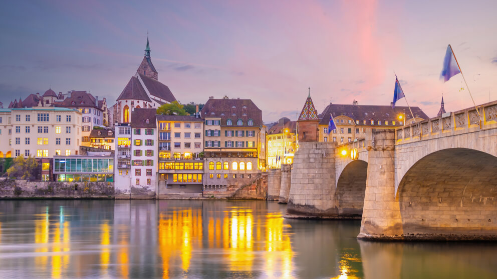 Basel am Flussufer bei Dämmerung mit beleuchteten Häusern, einer Kirche und einer historischen Steinbrücke mit Fahnen, Spiegelung im Wasser.