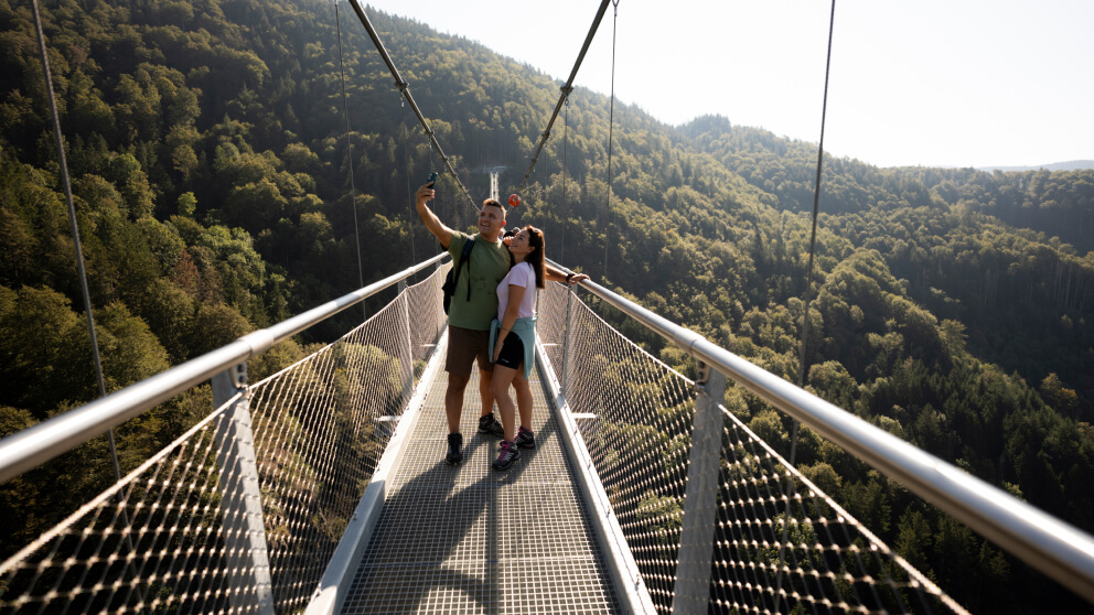 Paar macht ein Selfie auf der Blackforestline-Hängeseilbrücke hoch über einem bewaldeten Tal.