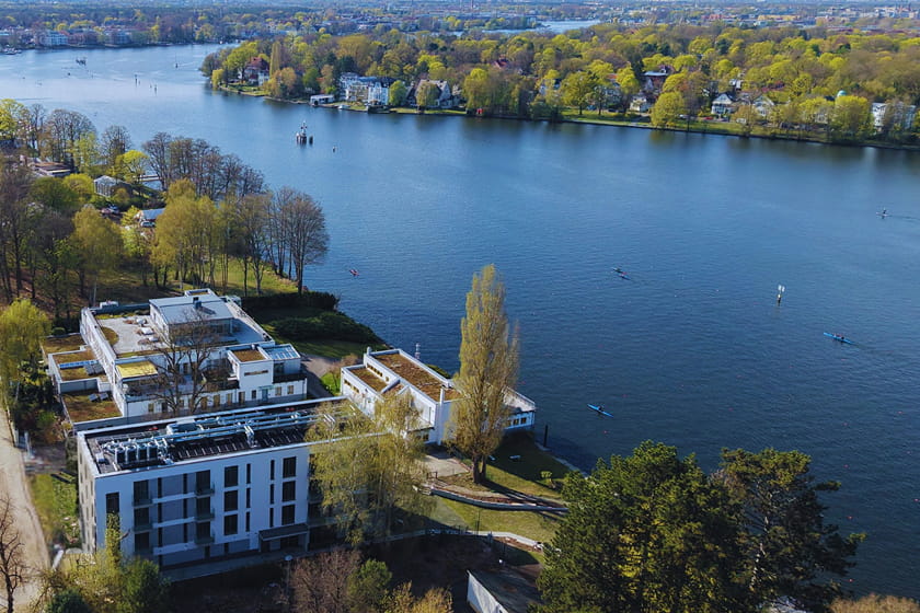Luftaufnahme moderner Gebäude an einem See, an dessen Ufer Menschen Kajak fahren und Bäume mit Frühlingslaub stehen.