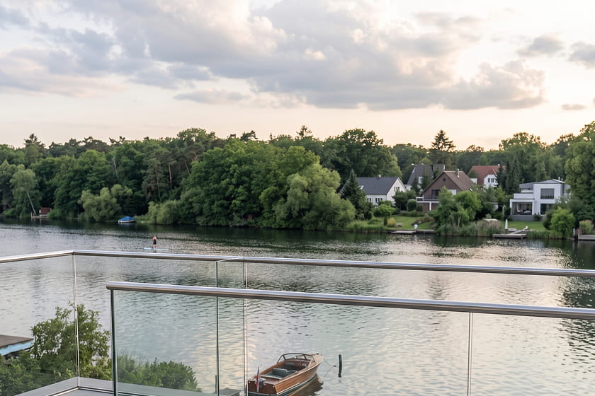 View from a balcony with a glass railing overlooking a river, a boat moored at the bank, and houses surrounded by trees on the opposite bank under a cloudy sky.