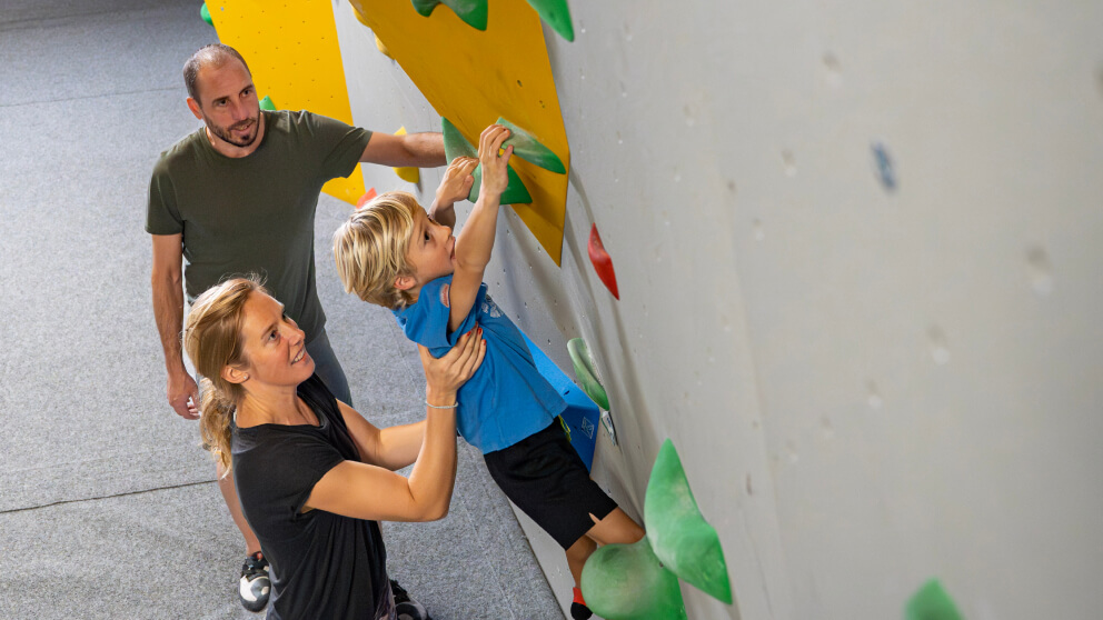 Junge klettert an einer Boulderwand, Vater und Mutter unterstützen ihn von unten in der Fundorena Kletterhalle.