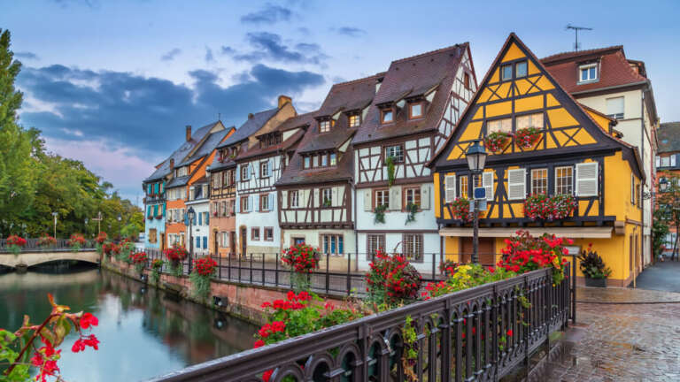 Bunte Fachwerkhäuser säumen einen Kanal, im Vordergrund ein Geländer mit roten Blumen, im Hintergrund eine kleine Brücke.
