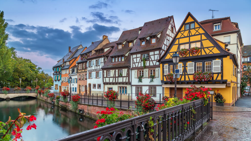 Bunte Fachwerkhäuser säumen einen Kanal, im Vordergrund ein Geländer mit roten Blumen, im Hintergrund eine kleine Brücke.