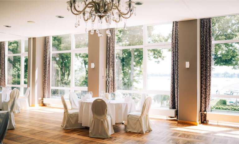 Eleganter Speisesaal mit Kristalllüster, weißen Stuhlhussen und großen Fenstern