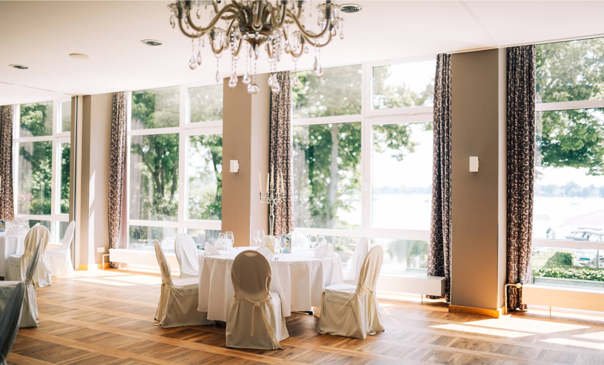 Eleganter Speisesaal mit Kristalllüster, weißen Stuhlhussen und großen Fenstern