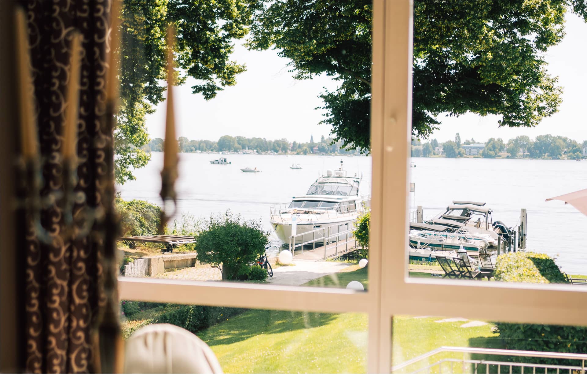 Fensterblick vom Restaurant auf Seeufer mit Booten und grüner Parklandschaft