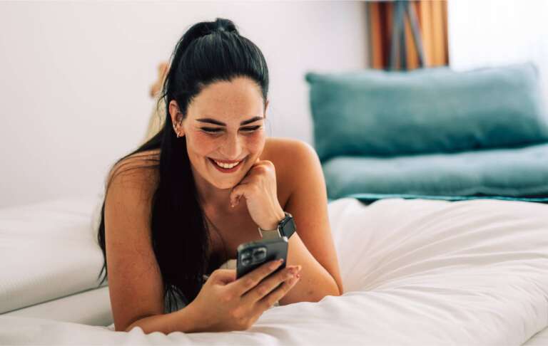 Lächelnde Frau mit Smartphone im Hotelbett, entspannte Atmosphäre