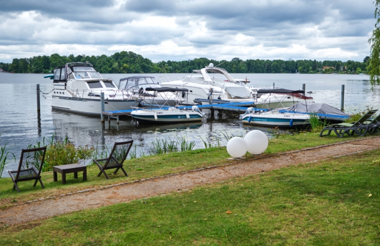 Mehrere Motorboote liegen an einer Anlegestelle am Dämeritzsee unter bedecktem Himmel. Im Vordergrund sind Stühle, ein kleiner Tisch und zwei große weiße Luftballons auf dem grasbewachsenen Ufer in der Nähe des Bootsverleihs Berlin und des H24 DämeritzSeehotels zu sehen.