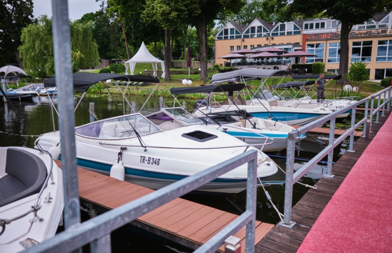 Mehrere weiß-blaue Boote liegen in einem Yachthafen in der Nähe des H24 DämeritzSeehotels. Eine Person steht auf dem Steg neben den Booten. Im Hintergrund sind Bäume und Gebäude zu sehen, die einen friedlichen Moment beim Bootsverleih Berlin einfangen.