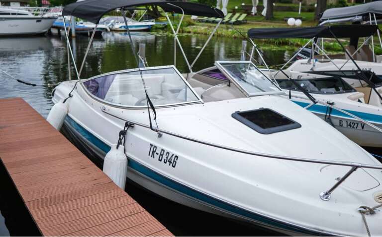 Mehrere Sportboote liegen vertäut an einem Steg auf einem ruhigen Gewässer. Im Vordergrund ist ein weißes Motorboot mit schwarzem Verdeck zu sehen. Die Umgebung wirkt gepflegt und einladend.