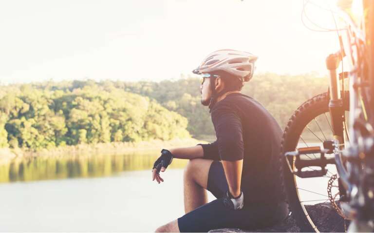 Ein Radfahrer mit Helm sitzt am Ufer eines Sees und blickt in die Ferne. Im Vordergrund steht sein Mountainbike, im Hintergrund sind bewaldete Hügel zu sehen.