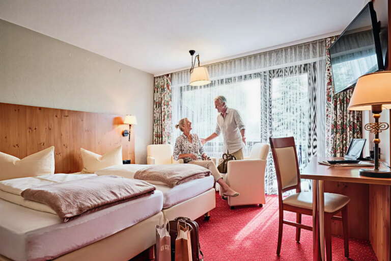 Ein älteres Ehepaar sitzt in einem hellen Zimmer des Hotel Hochfirst mit zwei Betten, einem Schreibtisch, Sesseln und einem großen Fenster mit durchsichtigen Vorhängen und lächelt sich an.