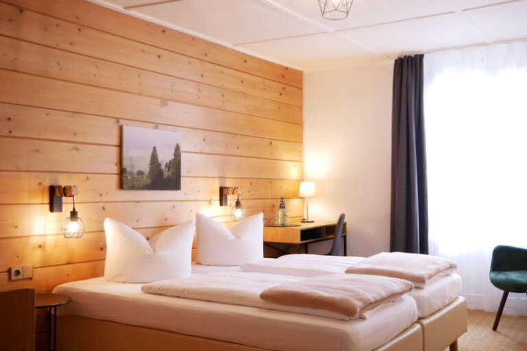 Ein ordentliches Hotel-Hochfirst-Zimmer mit einem Doppelbett, einer hölzernen Akzentwand, zwei Wandlampen, einem Schreibtisch mit Stuhl und einem Fenster mit Vorhängen, durch das Tageslicht fällt.