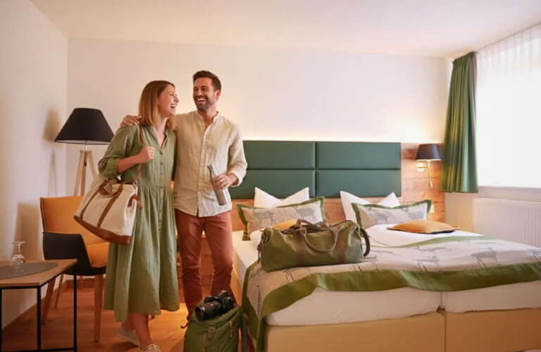 Ein lächelndes Paar steht neben einem sauber gemachten H24 Hotel Hochfirst-Bett mit Gepäck und scheint gerade in einem hellen, modernen Zimmer angekommen zu sein.