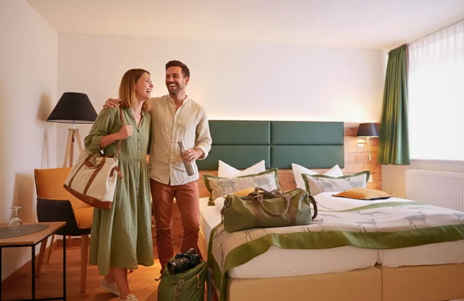 Ein lächelndes Paar steht neben einem sauber gemachten H24 Hotel Hochfirst-Bett mit Gepäck und scheint gerade in einem hellen, modernen Zimmer angekommen zu sein.