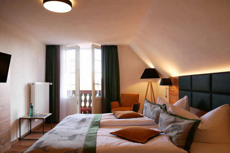 Ein ordentlich gemachtes Doppelbett mit grünen Akzenten in einem gemütlichen, modernen Hotel Hochfirst Zimmer mit Fenster, dunklen Vorhängen, Beistelltisch, Sessel und Stehlampe.