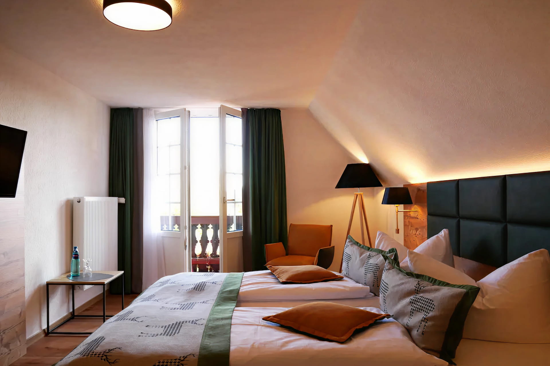 Ein ordentlich gemachtes Doppelbett mit grünen Akzenten in einem gemütlichen, modernen Hotel Hochfirst Zimmer mit Fenster, dunklen Vorhängen, Beistelltisch, Sessel und Stehlampe.