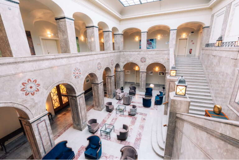 Geräumige, elegante Lobby mit Marmorsäulen, Bögen und Treppe; mehrere Sitzbereiche mit Sesseln und Tischen; natürliches Licht durch ein großes Oberlicht.