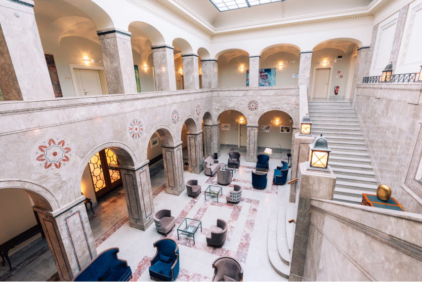 Geräumige, elegante Lobby mit Marmorsäulen, Bögen und Treppe; mehrere Sitzbereiche mit Sesseln und Tischen; natürliches Licht durch ein großes Oberlicht.