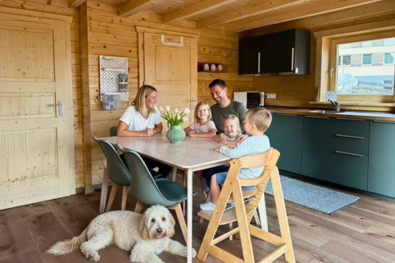 Eine fünfköpfige Familie sitzt um einen Esstisch in der gemütlichen Holzküche des Hotels Hochfirst und lächelt, während in der Nähe ein weißer Hund auf dem Boden liegt.