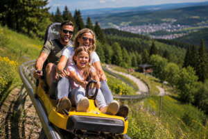 Drei Personen – zwei Erwachsene und ein Kind – fahren gemeinsam auf einer gelben Sommerrodelbahn eine Strecke den Hang hinunter, umgeben von Grün und mit Blick auf den Wald in der Ferne.