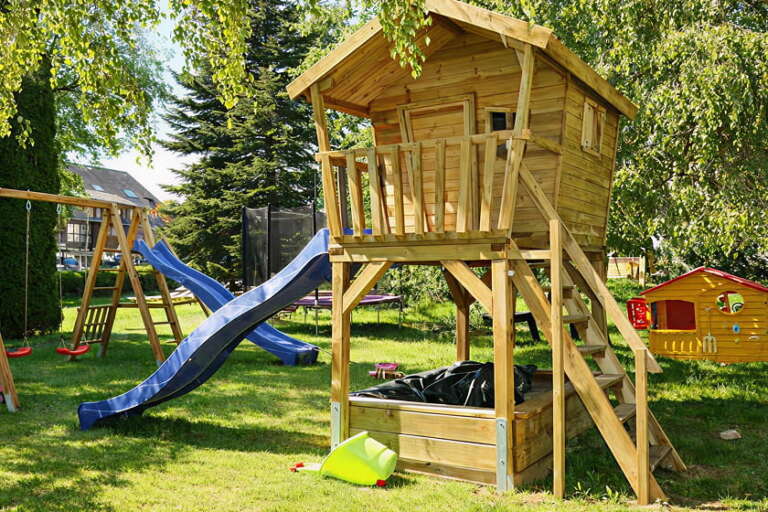 Ein Spielhaus aus Holz mit Balkon und Sandkasten, neben einer Rutsche, Schaukeln und einem weiteren kleinen Spielhaus in einem grasbewachsenen Garten.