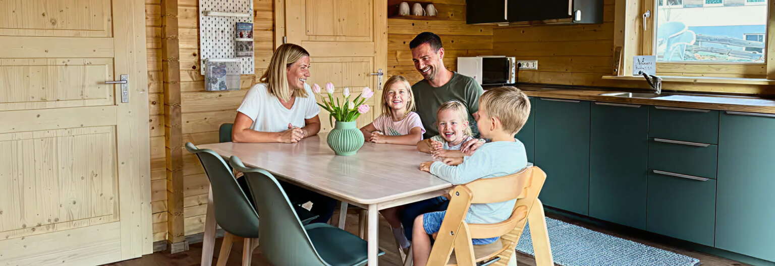 Eine fünfköpfige Familie sitzt um einen hölzernen Esstisch in einer modernen Küche mit grünen Schränken und holzvertäfelten Wänden, lächelt und unterhält sich miteinander.