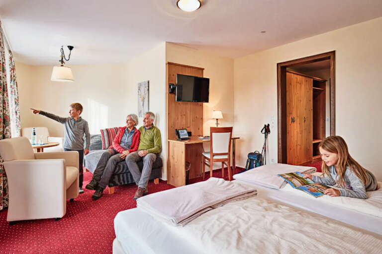 Ein Mädchen liest auf einem Bett eine Zeitschrift, während drei ältere Erwachsene in einem Zimmer des Hotels Hochfirst mit rotem Teppich und Holzmöbeln am Fenster sitzen und sich unterhalten.