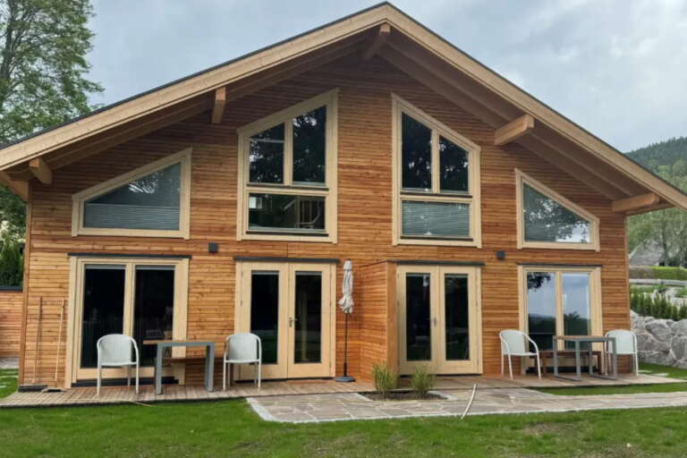 Eine moderne zweistöckige Blockhütte mit großen dreieckigen Fenstern, vier Glastüren, Außenstühlen und einer kleinen Rasenfläche davor - ganz im einladenden Stil des Hotels Hochfirst.
