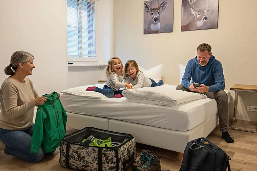 Eine Frau packt Kleidung aus, während zwei Kinder auf einem Bett lachen und ein Mann in der Nähe sitzt und auf seinem Handy tippt – in einem hellen, aufgeräumten Zimmer, an dessen Wänden Bilder von Rehen hängen.
