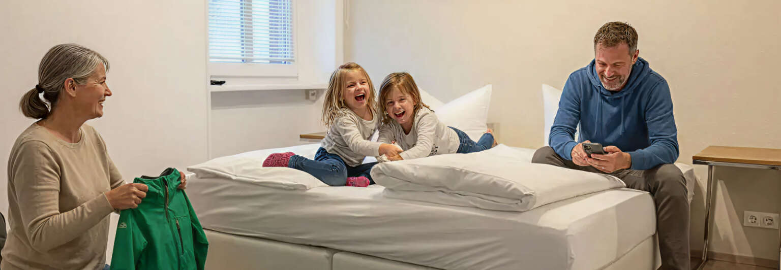 In einem schlichten, hellen Zimmer lachen zwei junge Mädchen auf einem Bett, während eine ältere Frau in der Nähe Wäsche zusammenlegt und ein Mann am Bettrand sitzt und auf sein Handy schaut.