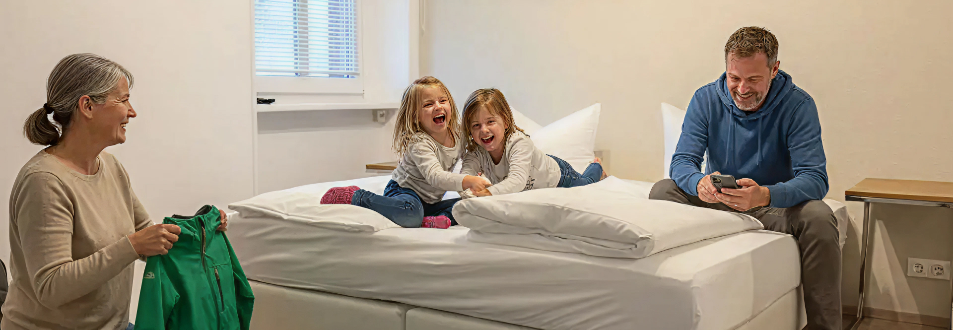 In einem schlichten, hellen Zimmer lachen zwei junge Mädchen auf einem Bett, während eine ältere Frau in der Nähe Wäsche zusammenlegt und ein Mann am Bettrand sitzt und auf sein Handy schaut.
