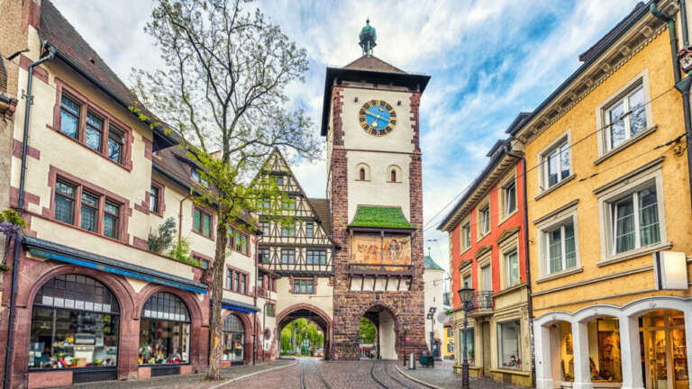 Historischer Stadttorturm mit Uhr in einer Altstadtgasse in Freiburg, umgeben von bunten Fachwerk- und Sandsteinhäusern mit Geschäften.