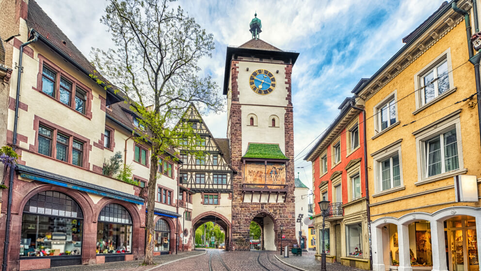 Historischer Stadttorturm mit Uhr in einer Altstadtgasse in Freiburg, umgeben von bunten Fachwerk- und Sandsteinhäusern mit Geschäften.