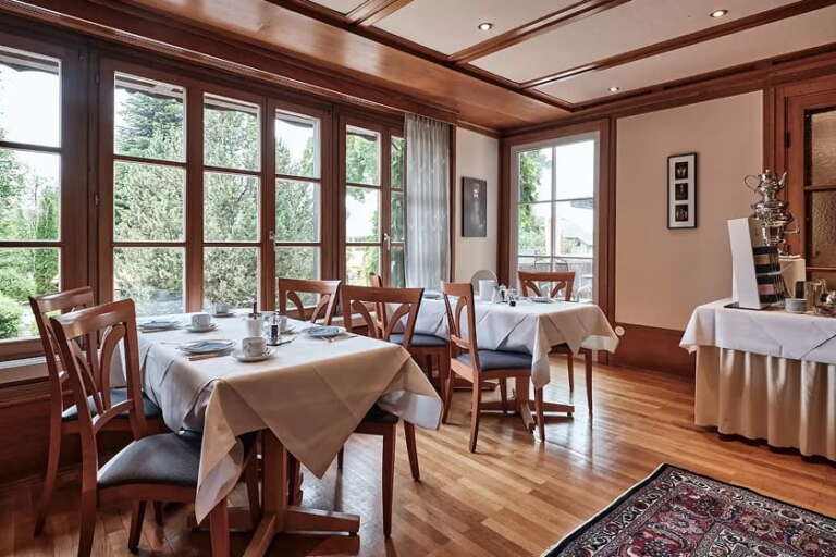 Ein Restaurantbereich mit Holzböden und großen Fenstern, in dem mehrere Tische mit weißen Tischdecken, Stühlen und Geschirr gedeckt sind.