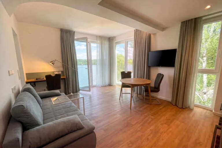 Modernes Wohnzimmer mit einem Sofa, einem Schreibtisch, einem runden Tisch, Stühlen, einem an der Wand montierten Fernseher, großen Fenstern und einem Blick auf Bäume und Wasser draußen.