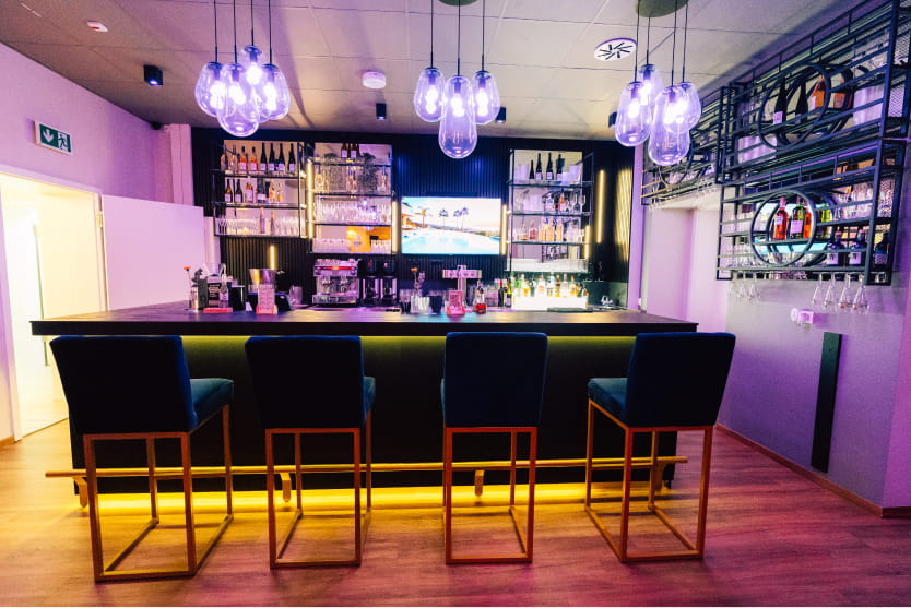 Eine moderne Bar im Parkhotel Thale mit vier Barhockern, beleuchteten Regalen mit Flaschen, Pendelleuchten über dem Kopf und einem Bildschirm hinter dem Tresen.