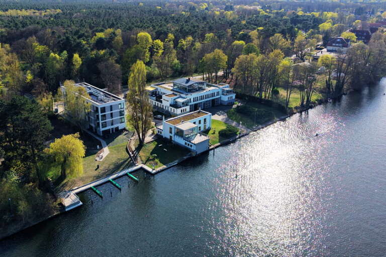 Luftaufnahme des Hotels H24 Berlin Grünau, umgeben von Bäumen und Grünflächen, mit Bootsanlegestellen entlang des Ufers und Sonnenlicht, das sich auf dem Wasser spiegelt.