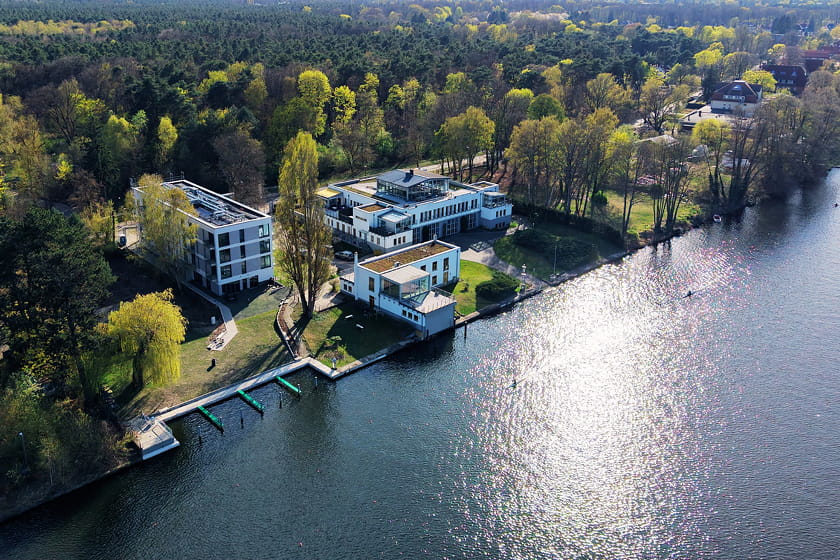 Luftaufnahme des Hotels H24 Berlin Grünau, umgeben von Bäumen und Grünflächen, mit Bootsanlegestellen entlang des Ufers und Sonnenlicht, das sich auf dem Wasser spiegelt.