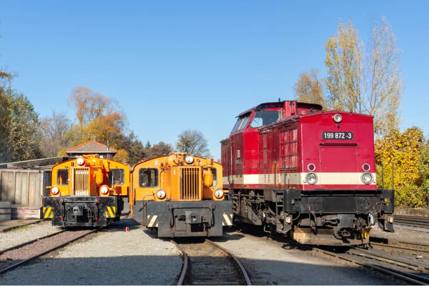 Historische Diesellokomotiven und eine rote Lok der Harzer Schmalspurbahnen auf Abstellgleisen im Bahnhofsumfeld
