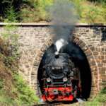 Schwarze Dampflokomotive der Harzer Schmalspurbahnen fährt aus einem steinernen Tunnel im grünen Mittelgebirge.