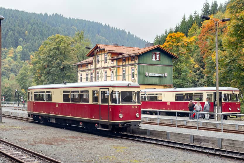 Zwei historische Triebwagen der Harzer Schmalspurbahnen stehen am Bahnhof vor bewaldeter Harz-Kulisse