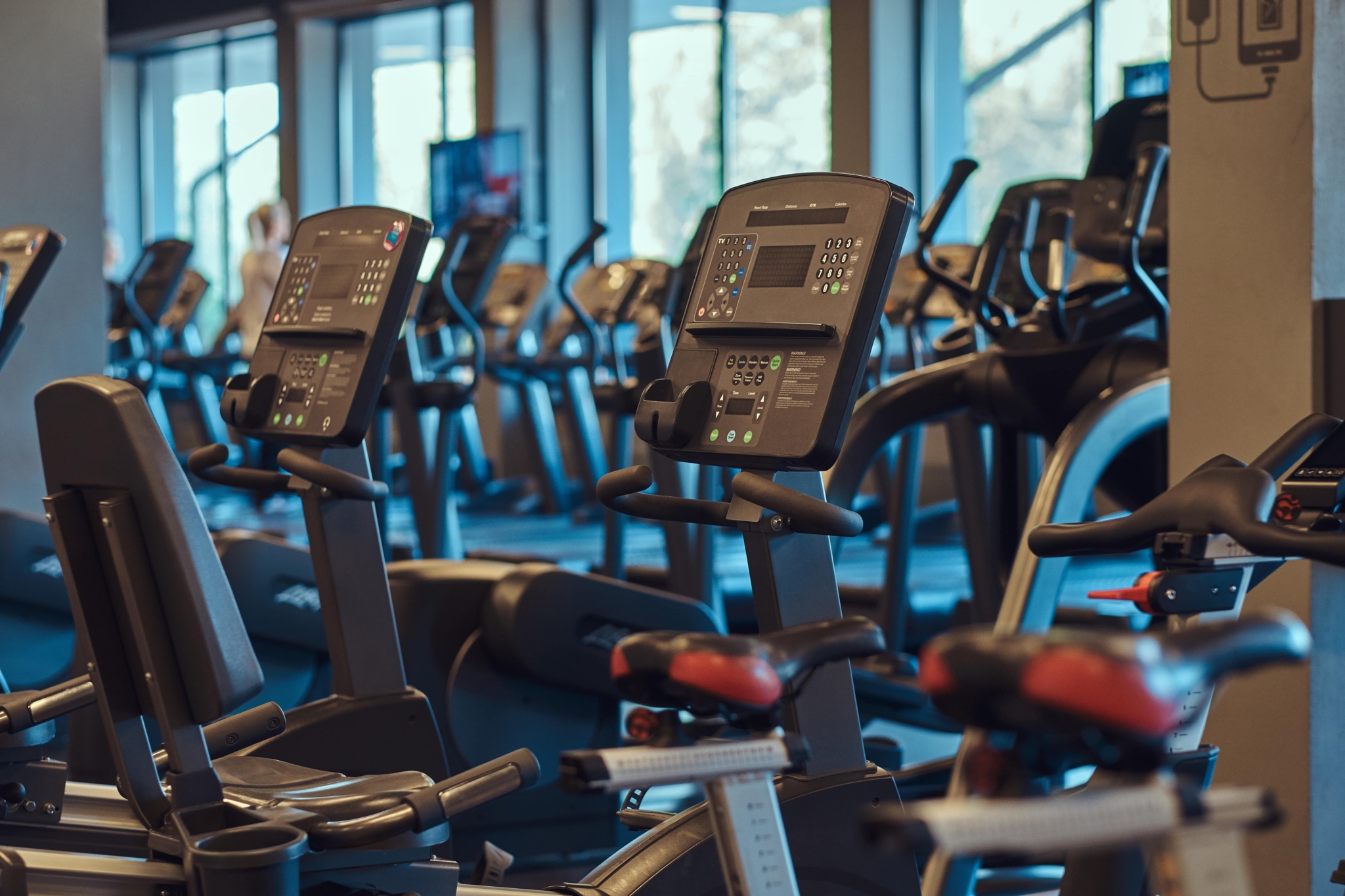 Eine Reihe stationärer Heimtrainer und Laufbänder in einem Fitnessstudio, mit großen Fenstern im Hintergrund, durch die natürliches Licht hereinfällt.