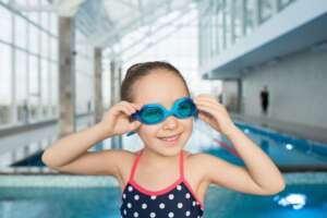 Ein junges Mädchen in einem gepunkteten Badeanzug rückt ihre blaue Schwimmbrille zurecht, während es neben einem Hallenbad steht.