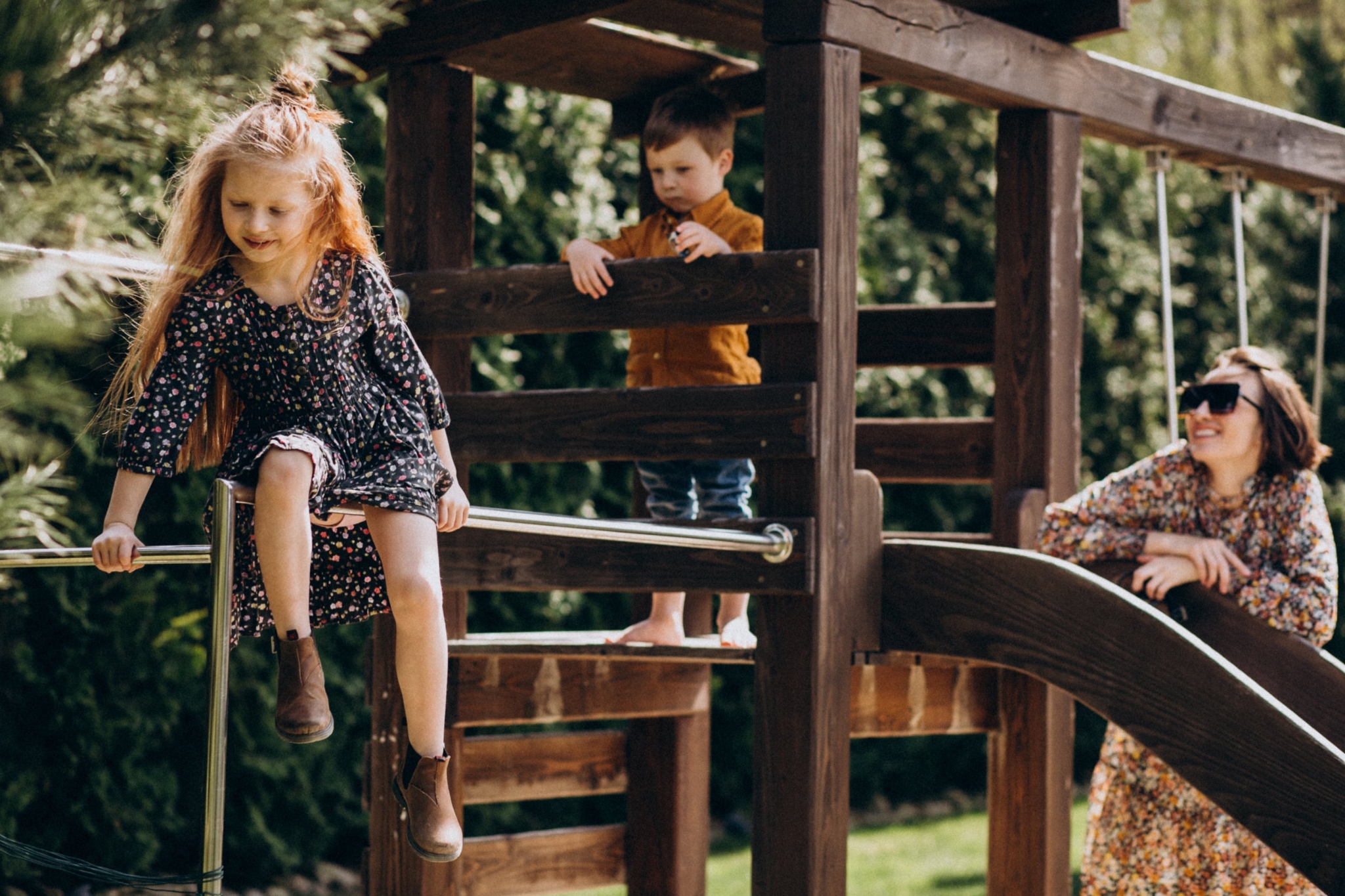 Ein junges Mädchen in einem geblümten Kleid sitzt auf der Stange eines Spielplatzgerüsts. Ein Junge in einem braunen Hemd steht auf dem Gerüst und eine Frau mit Sonnenbrille und geblümtem Kleid sieht ihnen lächelnd zu.